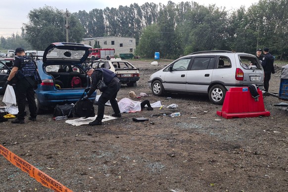 30.09.2022, Ukraine, Saporischschja: Eine Leiche liegt abgedeckt nach einem Raketenangriff auf dem Boden, während Polizisten an Autos Gepäck untersuchen. Bei einem Raketenangriff auf einen zivilen Aut ...