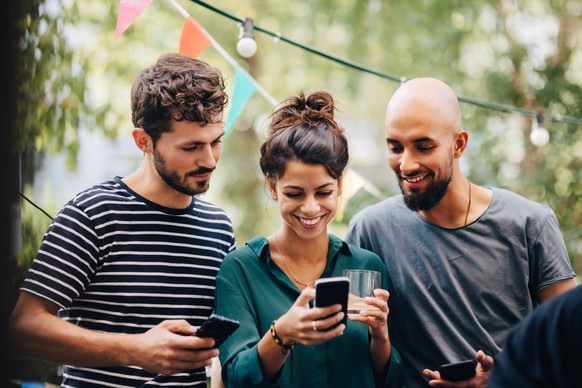 Happy young woman showing mobile phone to male friends while standing in balcony during party , model released, property released Copyright: xMaskotx , MA68073