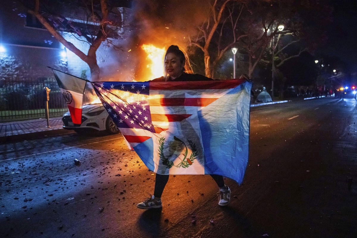 A person displaying multiple flags walks past a burning car during protests over the Trump administration&#039;s immigration raids in Los Angeles, Monday, June 9, 2025. (AP Photo/Ethan Swope)