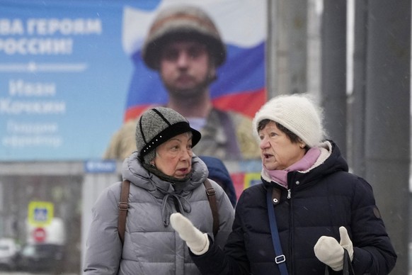 31.01.2023, Russland, St. Petersburg: Frauen unterhalten sich beim Vorbeigehen an einem Plakat mit dem Porträt eines russischen Soldaten, der für seinen Einsatz in der Ukraine ausgezeichnet wurde, und ...