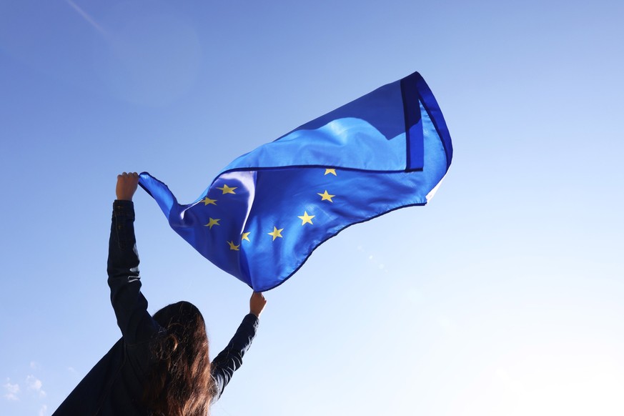Woman holding European Union flag against blue sky outdoors, low angle view Model Released Property Released xkwx angle back background blue brussels campaign clear cloth color community concept coope ...