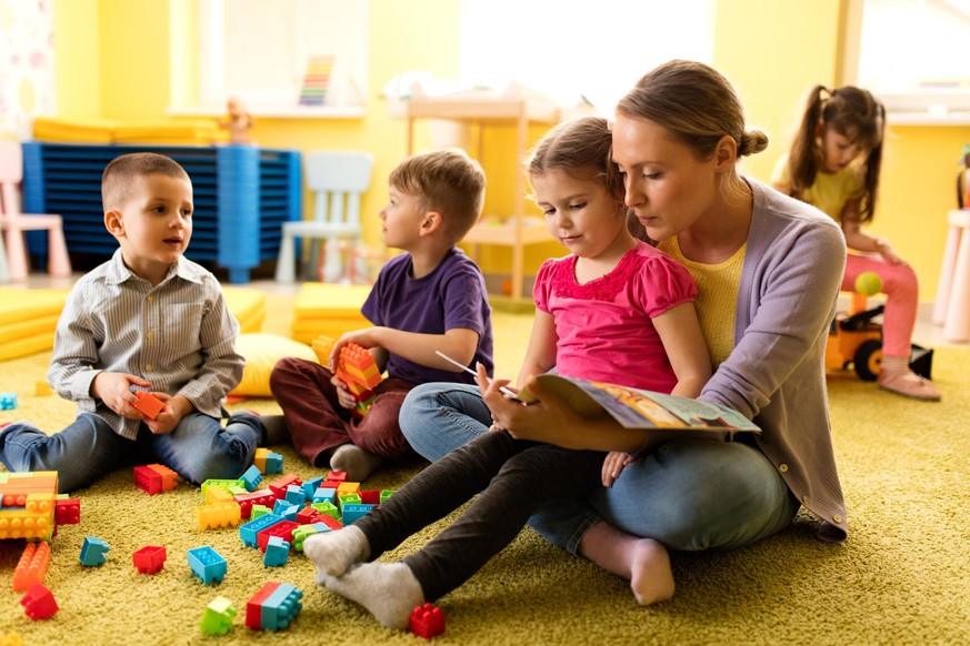 Cute little girl sitting in teacher&#039;s lap in kindergarten and reading a story while the other children are playing.