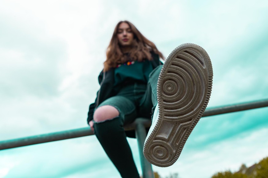 Teenage Girl sitting on fence stretching out her leg, showing the sport shoe rubber sole. Teenage Lifestyle Portrait. Shot from below, Selective Focus on Sport Shoue Rubber Sole.