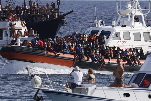 18.09.2023, Italien, Lampedusa: Ein Boot der italienischen Küstenwache transportiert Migranten unter der Beobachtung einer Gruppe von Touristen. Die Ankunft Tausender Bootsmigranten in kurzer Zeit hat ...