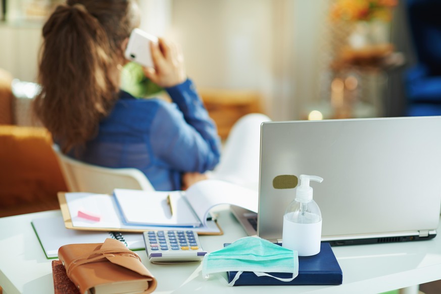 Closeup on medical mask and hand disinfectant and woman in background talking on smartphone in temporary home office during the coronavirus epidemic at home in sunny day.