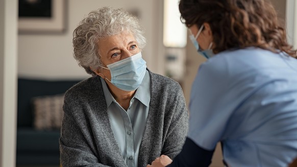 Elderly woman talking with a doctor while holding hands at home and wearing face protective mask. Worried senior woman talking to her general pratictioner visiting her at home during virus epidemic. D ...