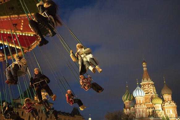 25.12.2022, Russland, Moskau: Kinder und ihre Eltern fahren auf einem Karussell auf dem Weihnachtsmarkt auf dem Roten Platz, der für die Neujahrs- und Weihnachtsfeierlichkeiten geschmückt ist. Im Hint ...