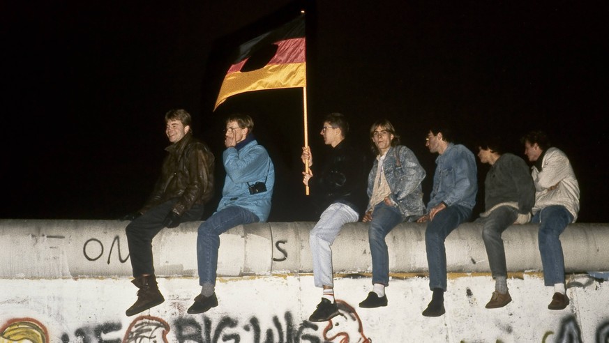 Berliner feiern ohne das ausgeschnittene Emblem aus der DDR-Fahne den Fall der Berliner Mauer rittlings auf eben jener sitzend im nächtlichen Berlin.