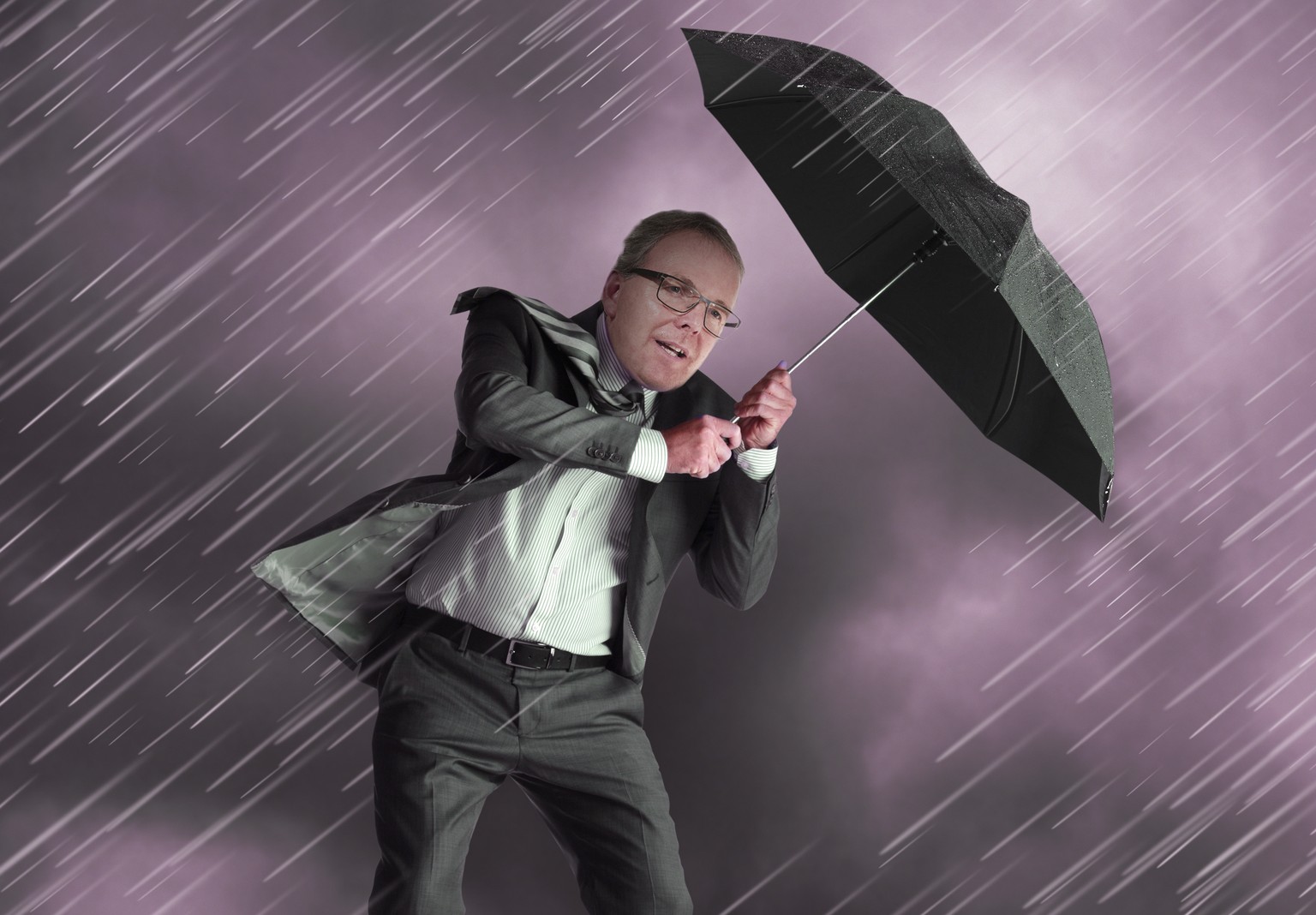 A businessman with an umbrella protecting himself from a driving rainstorm while standing close to the edge of a cliff.