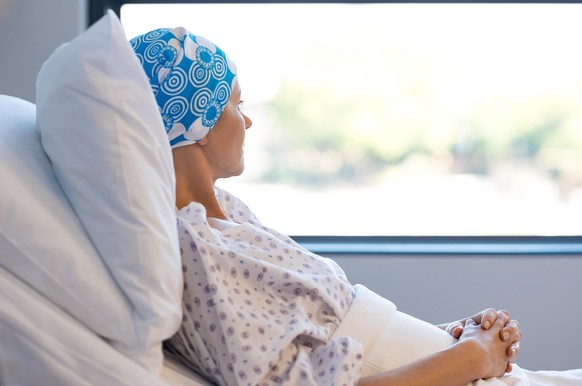 Young woman in bed suffering from cancer. Thoughtful woman battling with tumor looking out of window. Young patient with blue headscarf recovery in hospital on bed.