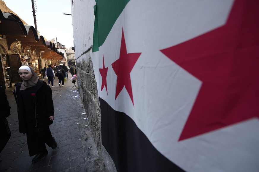 21.12.2024, Syrien, Damaskus: Eine Frau geht an der syrischen &quot;Revolutions&quot;-Flagge vorbei, die an einer Wand in der Altstadt von Damaskus, Syrien hängt. Foto: Hussein Malla/AP/dpa +++ dpa-Bi ...