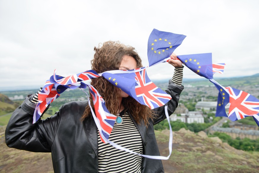 Brexit - Young adult woman holding European Union and British Union Jack flags on a windy hilltop. Arthur's Seat, Edinburgh, Scotland.