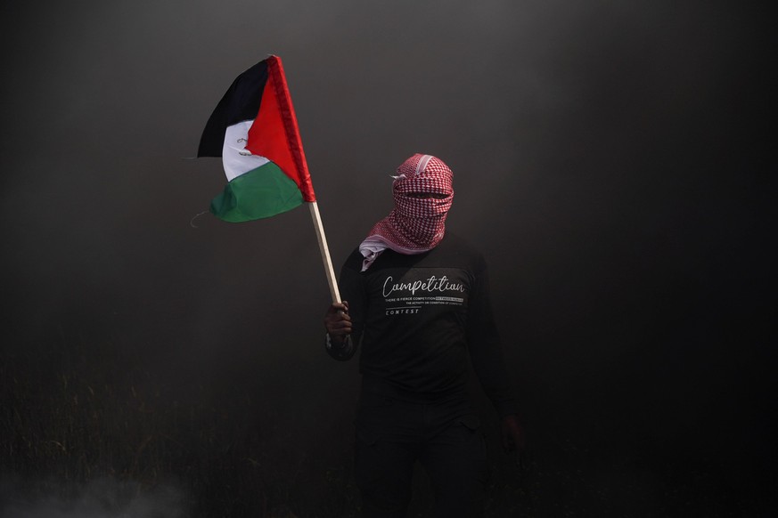 19.03.2023, Palästinensische Gebiete, Gaza: Eine Person steht im Rauch und hält eine palästinensische Flagge in der Hand. Palästinenser zündeten Gummireifen an, um ihren Unmut gegenüber des Gipfels zw ...
