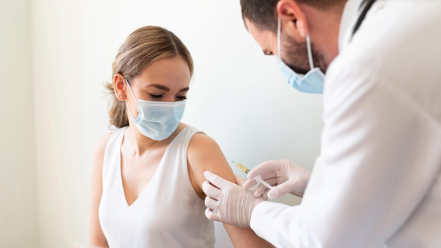 Closeup of a nervous woman and her doctor wearing face masks and getting a vaccine shot in a doctor&#039;s office