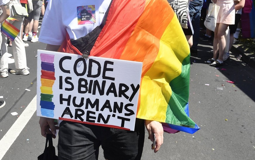 ARCHIV - 03.07.2022, Nordrhein-Westfalen, Köln: Menschen feiern bei der diesjährigen CSD-Parade - ein Teilnehmer hält ein Plakat mit der Aufschrift &quot;Code is binary - humans aren&#039;t&quot;. Per ...