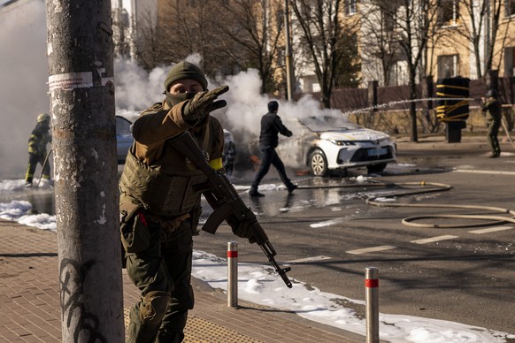 26.02.2022, Ukraine, Kiew: Ukrainische Soldaten beziehen Stellung vor einer Milit�reinrichtung, w�hrend Feuerwehrleute zwei brennende Autos l�schen. Russische Truppen haben den erwarteten Angriff auf  ...