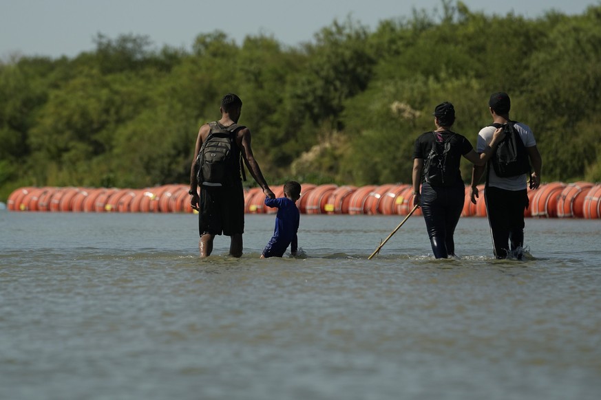 01.08.2023, USA, Eagle Pass: Migranten, die von Mexiko in die USA einreisen, gehen im Fluss Rio Grande an großen Bojen entlang, die als schwimmende Grenzbarriere eingesetzt werden. Foto: Eric Gay/AP + ...