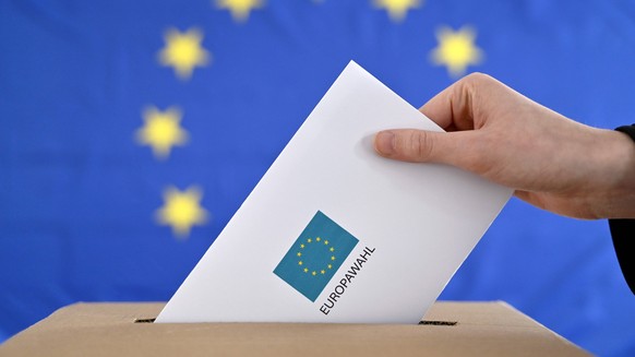 Ein Wahlbrief wird in eine Wahlurne gesteckt, im Hintergrund eine EU-Flagge. Die Europawahl ist eine seit 1979 in der Europäischen Union alle fünf Jahre stattfindende unmittelbare, freie und geheime W ...