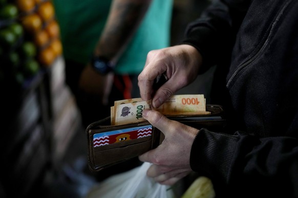 ARCHIV - 01.09.2023, Argentinien, Buenos Aires: Ein Mann holt mehrere 1000-Peso-Geldscheine aus seinem Geldbeutel vor einem Gemüsestand auf einem Markt. Die Argentinier haben mit einer der höchsten In ...