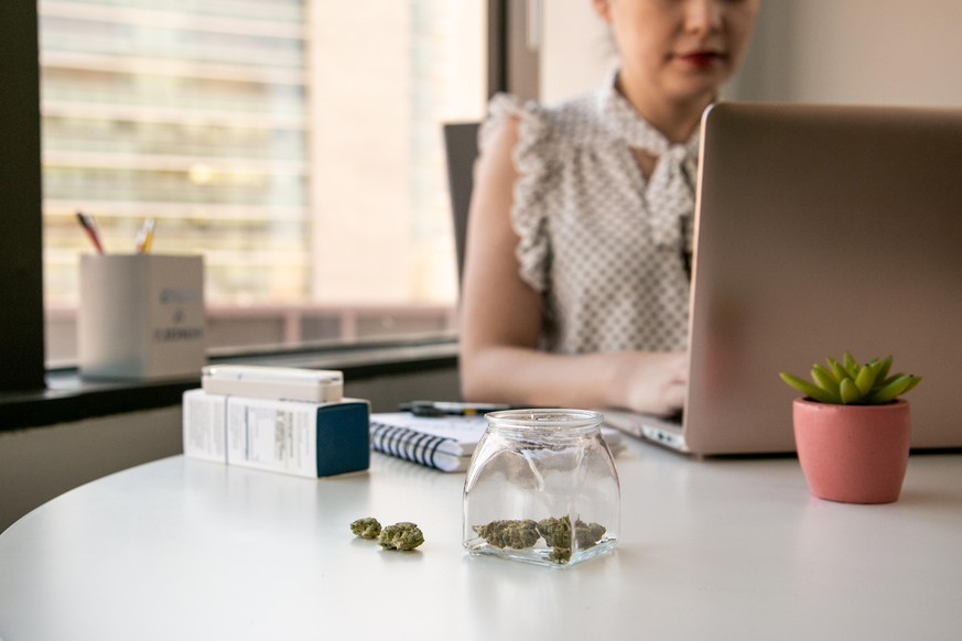 Glass jars of cannabis and buds sit on a white office table in an office environment with a downtown view outside the window. A Female Entrepreneur is working on a laptop computer with a vape pen by h ...