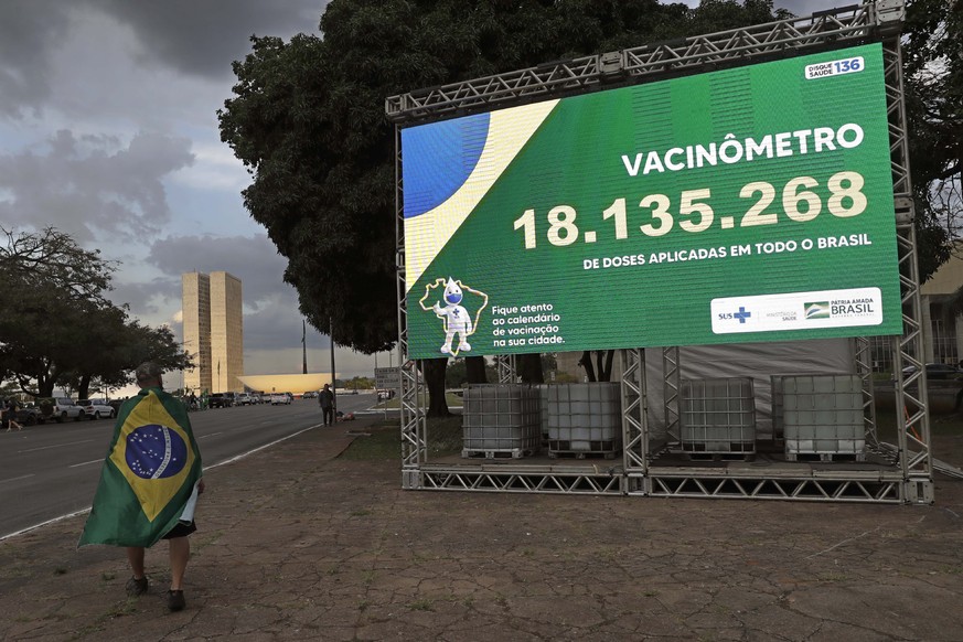 30.03.2021, Brasilien, Brasilia: Ein Mann mit einer brasilianischen Nationalflagge geht an einer Plakatwand mit der Zahl der verabreichten Covid-19-Impfstoffdosen vorbei. Inmitten einer Regierungskris ...