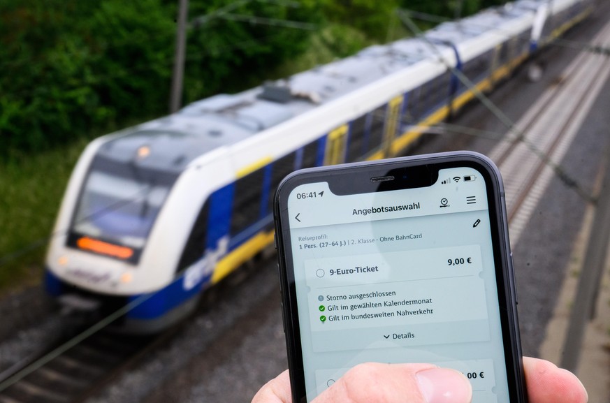 Auf einem Smartphonedisplay ist die Bahn-App DB Navigator zu sehen, in der man das 9-Euro-Ticket digital kaufen kann, während im Hintergrund ein Regionalzug vorbeifährt (gestellte Szene). Die Deutsche ...