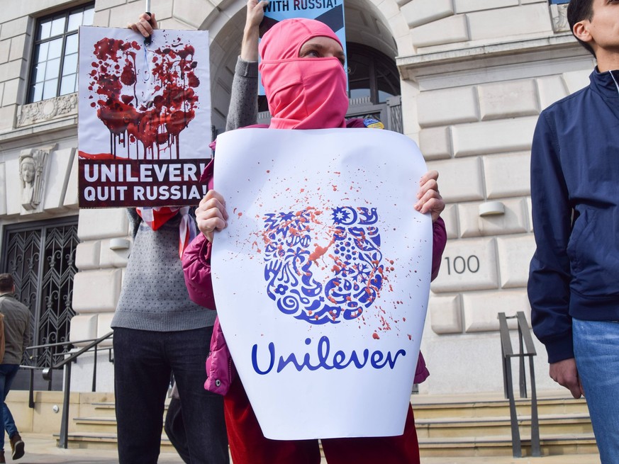 March 15, 2022, London, England, United Kingdom: A protester wearing a balaclava holds a sign with the Unilever logo splattered with fake blood. Protesters gathered outside the Unilever headquarters a ...