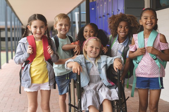 Front view of happy school kids standing in outside corridor at school while a Caucasian schoolgirl is sitting on wheelchair in foreground