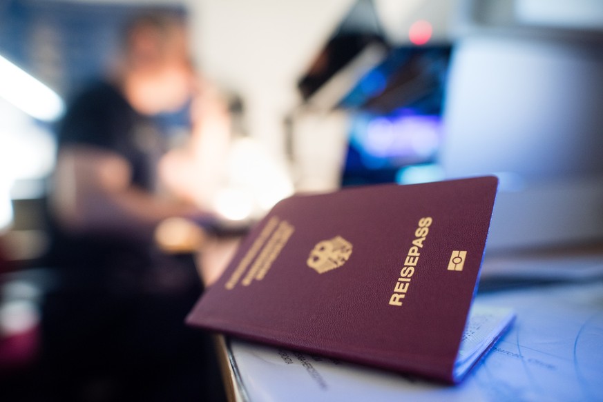 ARCHIV - 04.07.2016, Bayern, München: Ein deutscher Reisepass liegt in einem Labor der Kriminaltechnik der Bundespolizei am Flughafen, während im Hintergrund ein Urkundenprüfer der Bundespolizei an ei ...