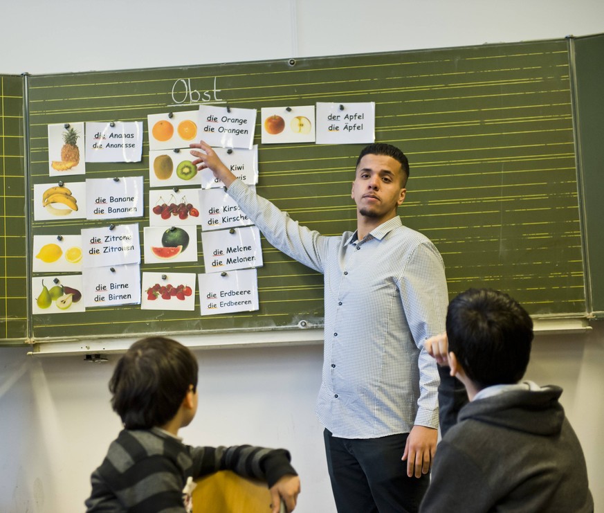 Teilnehmer am Bundesfreiwilligendienst ( Bufdi ) Mohamed Rahal, vor zwei Schuelern aus Syrien an der Tafel beim Deutschunterricht in einer Willkommensklasse fuer Migranten an der Leo-Lionni-Schule. Be ...