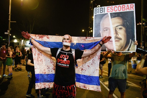 10.08.2024, Israel, Tel Aviv: Ein geknebelter Demonstrant mit falschem Blut an den H�nden und einer mit falschem Blut verschmierten israelischen Flagge steht neben einem Plakat mit der Aufschrift &quo ...