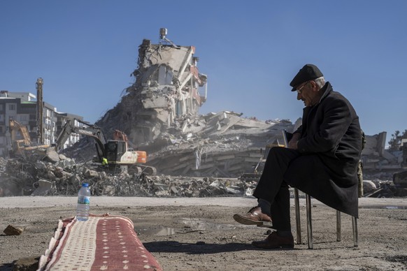 09.02.2023, Türkei, Nurdagi: Der 67-jährige Mehmet sitzt auf einem Stuhl, während im Hintergrund Bagger Trümmer aus einem Gebäude entfernen, in dem fünf seiner Familienmitglieder verschüttet sind. 
Di ...
