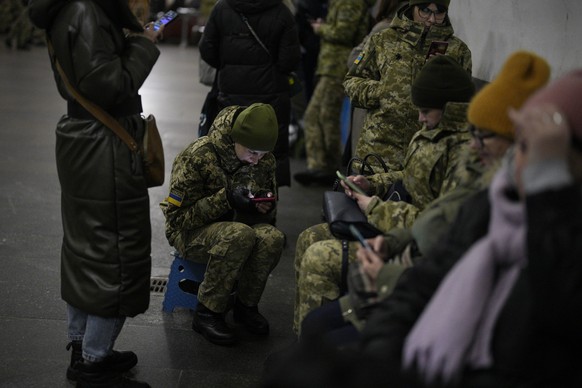 03.02.2023, Ukraine, Kiew: Soldatinnen und Soldaten schauen auf ihr Handy während sie mit Menschen in einer U-Bahn-Station, die als Luftschutzbunker genutzt wird, während einer Luftschutzsirene Schutz ...