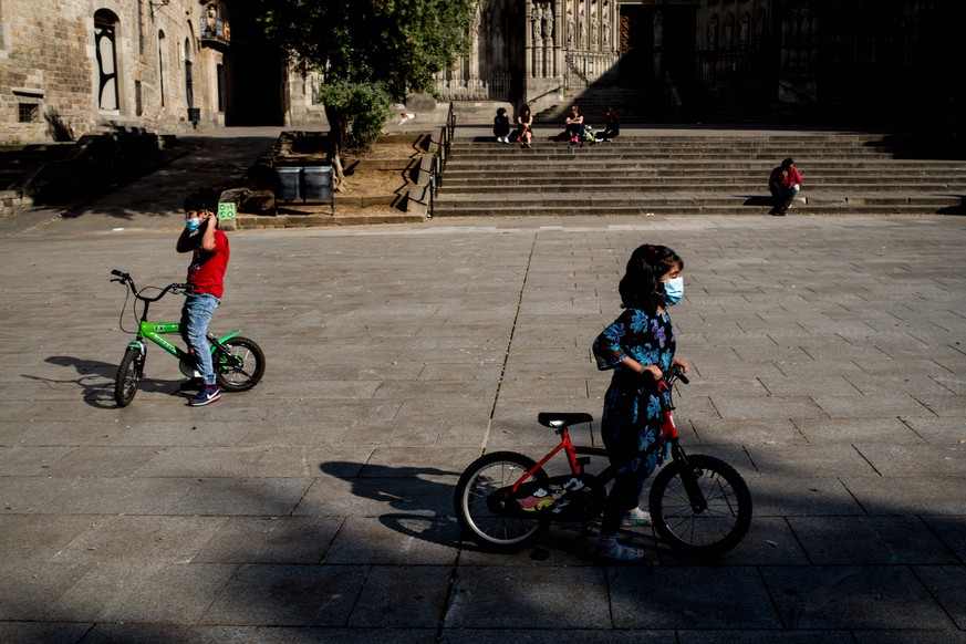 April 26, 2020, Barcelona, Catalonia, Spain: Children with protective face masks riding a bicycle in Barcelona s Cathedral square. In Spain from this Sunday 26 April and after 6 weeks of confinement,  ...