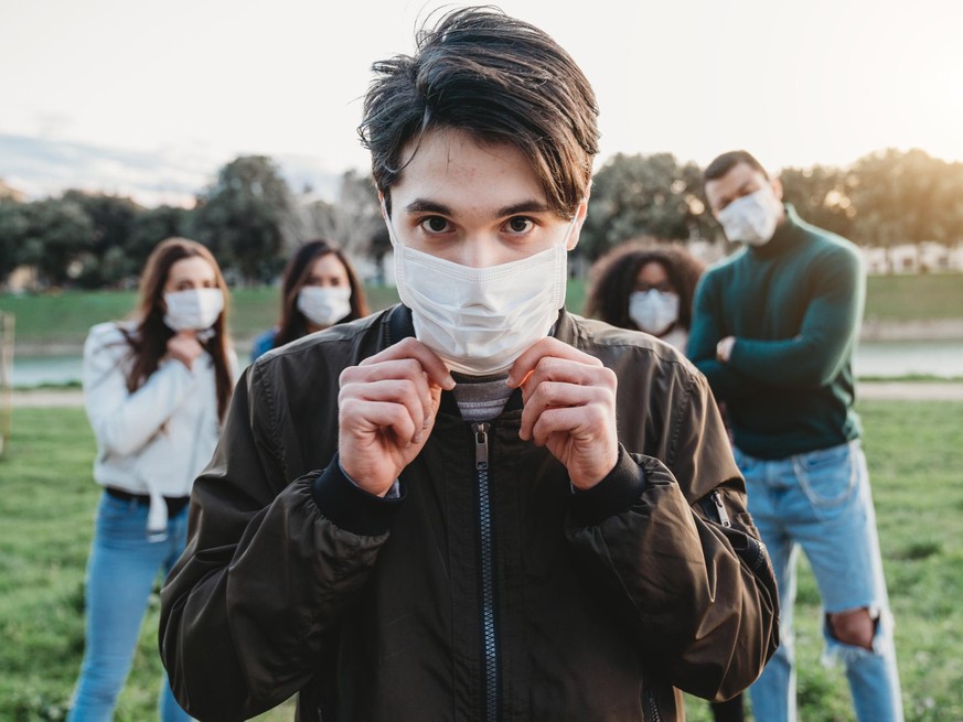 Young adult man wearing a pollution mask to protect himself from viruses. His friends are in the background. They all are wearing masks. Conceptual image of protection against pandemic virus.