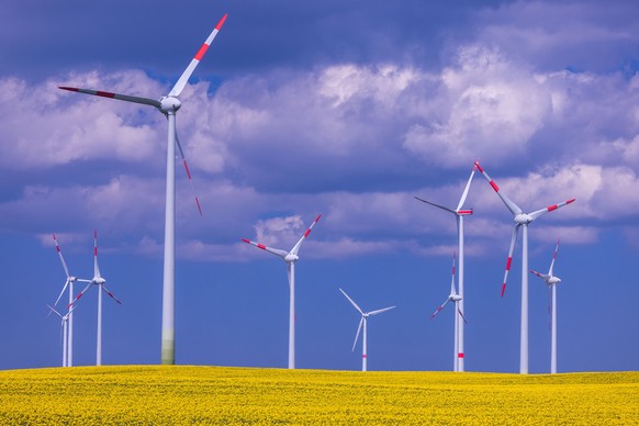 ARCHIV - 26.04.2022, Mecklenburg-Vorpommern, Kladrum: Windkraftanlagen verschiedener Hersteller drehen sich hinter einem blühenden Rapsfeld in einem Windpark. Um den Bau von Erneuerbaren Energien zu b ...