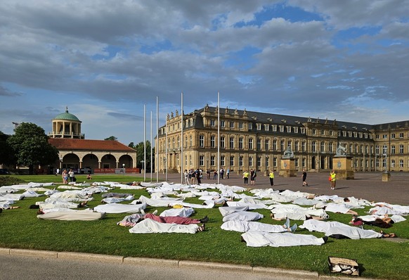 20.06.2023, Baden-Württemberg, Stuttgart: Menschen liegen mit weißen Laken zugedeckt auf der Wiese auf dem Schlossplatz. Bei der Protestaktion haben sich den Initiatoren zufolge rund 200 Menschen auf  ...