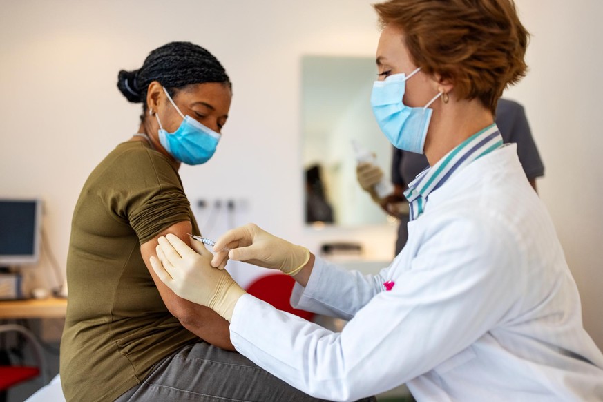 Female doctor wearing a face mask giving vaccine to a mature patient in her clinic. General practitioner giving flu shot to a senior woman in clinic.