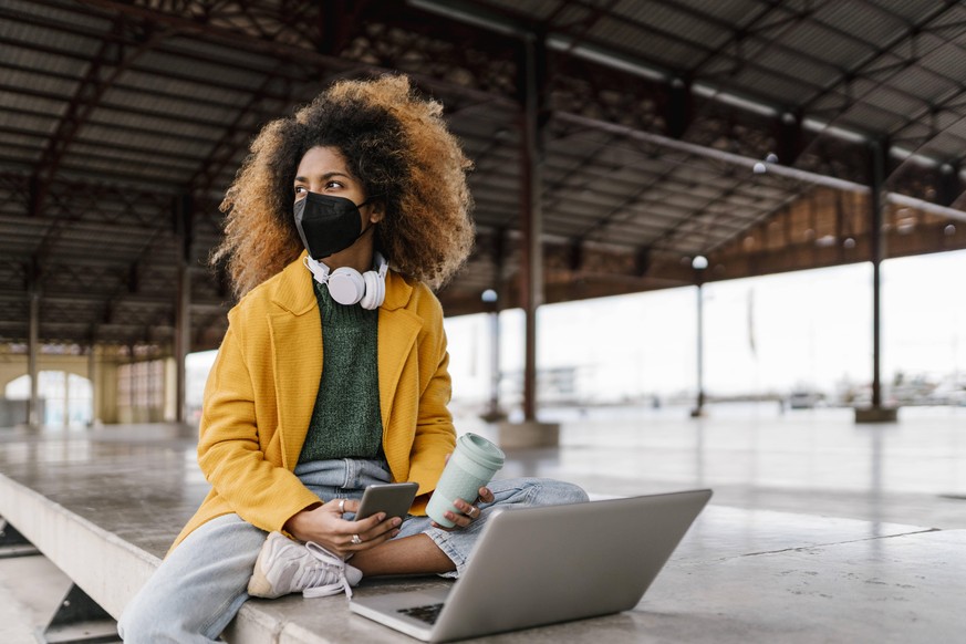 Afro woman with mobile phone and coffee cup looking away while sitting with laptop at seating area during pandemic model released Symbolfoto EGAF01994