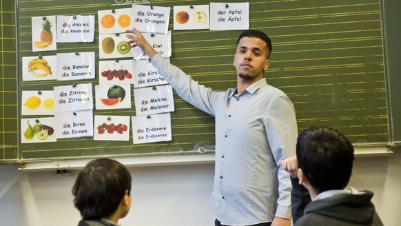 Teilnehmer am Bundesfreiwilligendienst ( Bufdi ) Mohamed Rahal, vor zwei Schuelern aus Syrien an der Tafel beim Deutschunterricht in einer Willkommensklasse fuer Migranten an der Leo-Lionni-Schule. Be ...