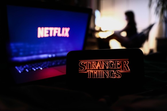Popular Netflix Original Series Stranger Things series logo displayed on a phone screen, Netflix logo displayed on a laptop screen and a silhouette of a woman in the background are seen in this illust ...