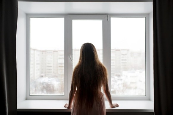 Rear view of woman silhouette standing near window looking out, melancholy and crisis concept