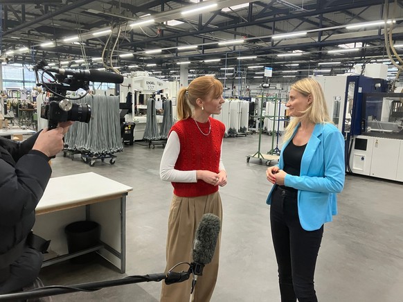 Eva Schulz steht in einer großen Halle und interviewt eine Protagonistin.