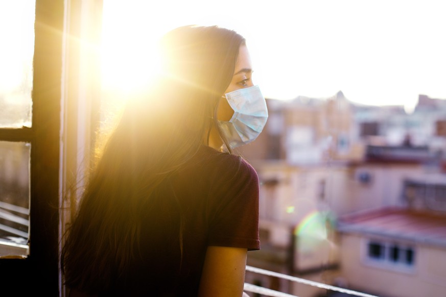 teenage girl wearing protective mask, looking at the city from the window during coronavirus qurantine in Barcelona. Nice sunset with the sun shining in the sky