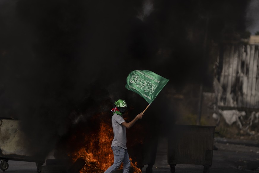 A Palestinian protester with a Hamas head band carries a Hamas flag behind burning tires during clashes following a demonstration in solidarity with the Gaza Strip, in the West Bank city of Ramallah,  ...