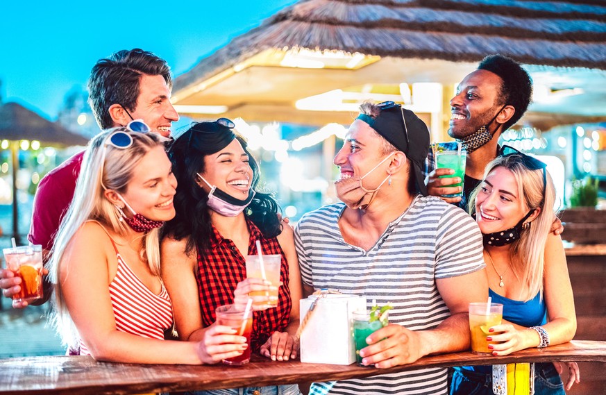 Young international friends talking at beach cocktail bar with open face mask - New normal tourism concept with millennial people having fun together - High iso image with focus on guy with black cap