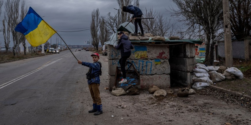 23.11.2022, Ukraine, Cherson: Ukrainische Kinder spielen und schwenken eine ukrainische Fahne an einem verlassenen Kontrollpunkt. Neue russische Raketenangriffe haben zu großflächigen Blackouts in der ...