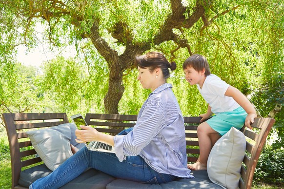 Alleinerziehende Mutter arbeitet am Laptop PC im Garten mit neugierigem Sohn dabei || Modellfreigabe vorhanden