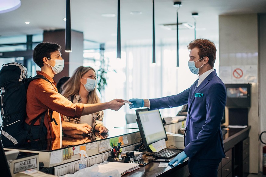 Three people, young couple travelers with medical masks on hotel reception talking to male receptionist.