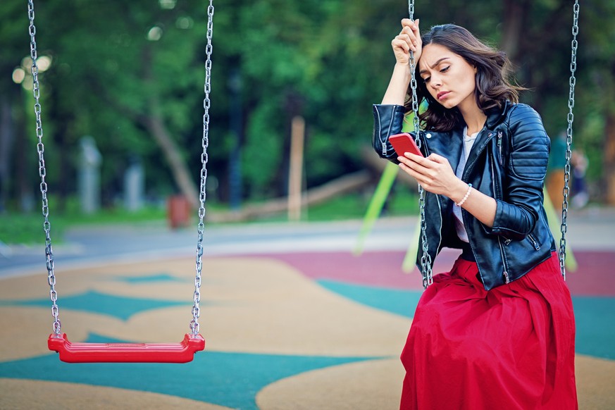 Sad young girl is sitting on the swind and looking her mobile phone